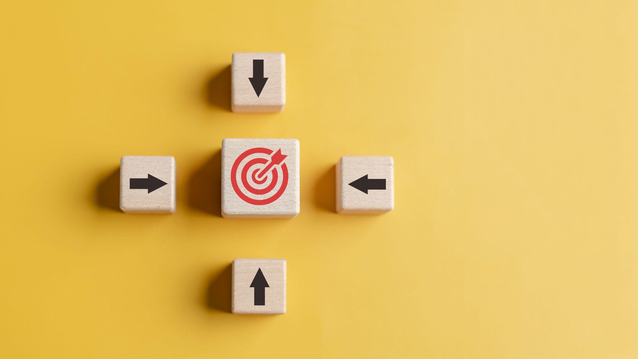 Five wooden blocks on a yellow background. Four blocks printed with arrows point inward from above, below, left and right at a central block showing a red target with an arrow in the bullseye.