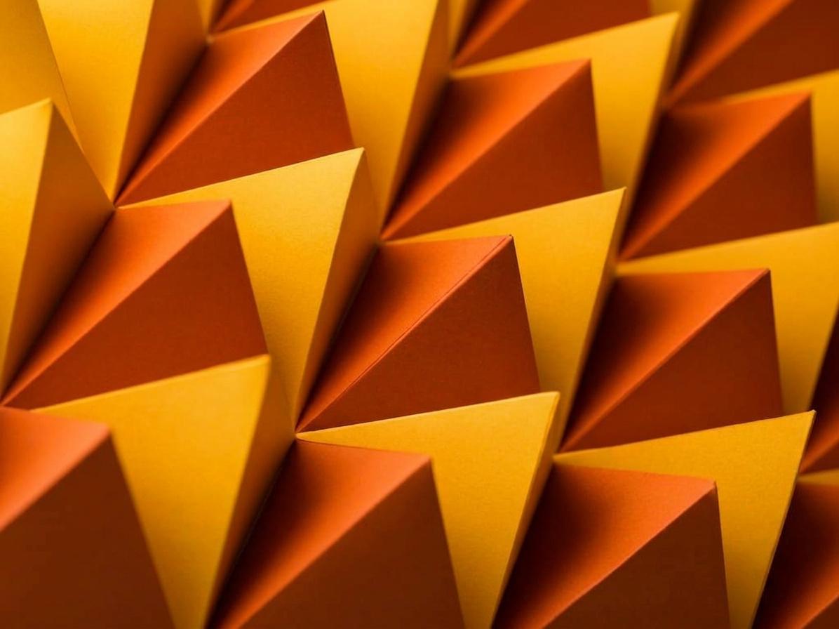 A close-up of folded paper arranged as a repeating grid of three-sided pyramids, lit so each pyramid shows one yellow face and one darker orange face.